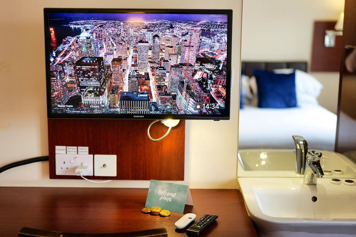 Guest room with a wall-mounted Smart TV above a wooden desk, a basin and a bed with navy cushions
